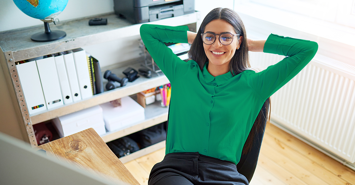 A person wearing glasses and a green shirt is sitting at a desk with hands behind their head, smiling. There's a bookshelf with folders and a globe in the background, and sunlight coming through a window.