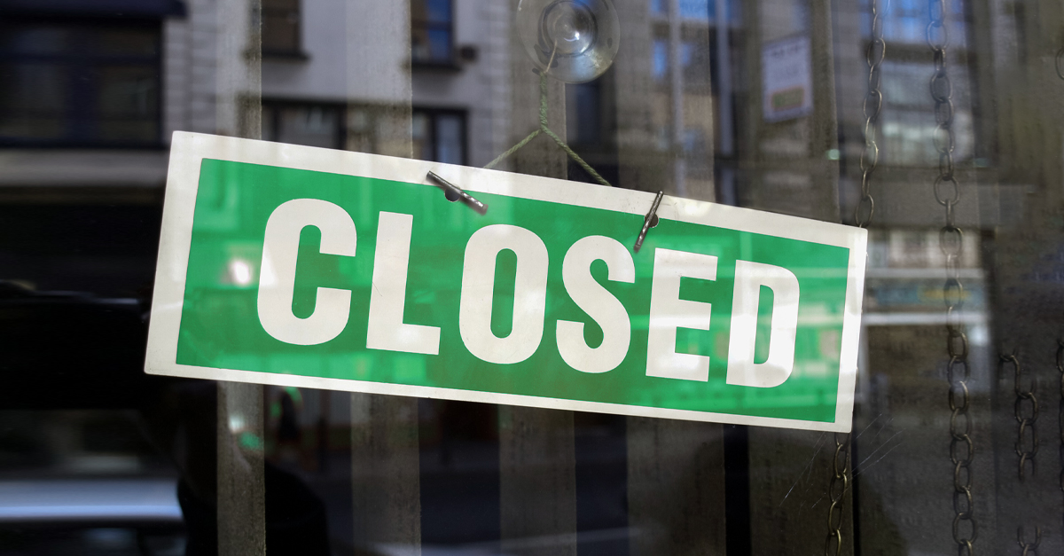 A green sign with white letters reading "CLOSED" hangs on a glass door, a subtle reminder of business failure. The reflection shows blurry buildings and windows, indicating an urban setting.