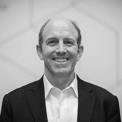 Smiling man in a suit jacket and collared shirt against a light geometric background, in black and white.