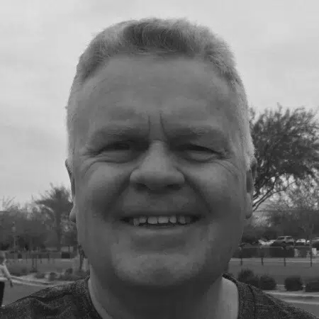 Smiling man with short hair outdoors, trees and cloudy sky in the background, black and white photo.